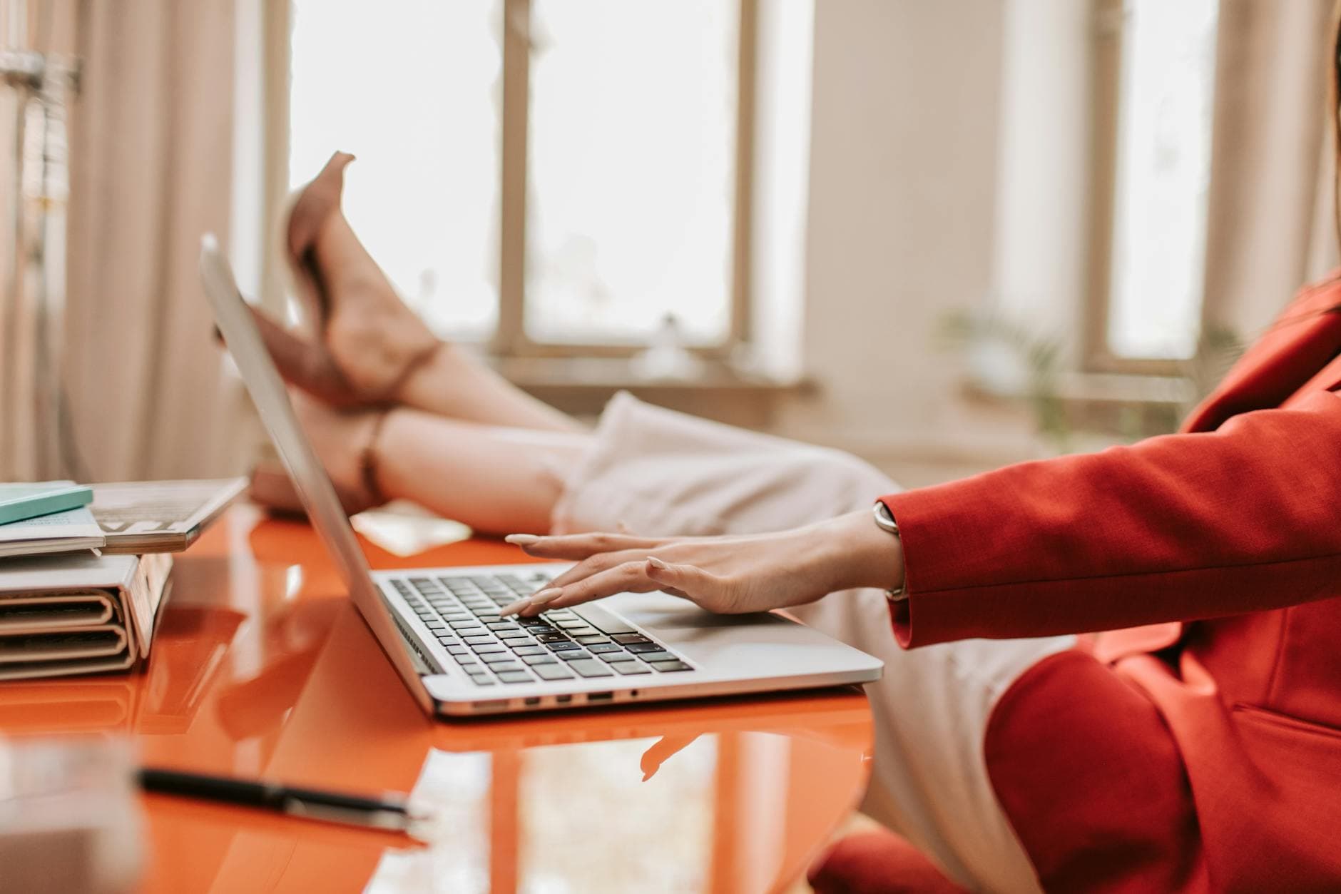 Businesswoman in red blazer working on laptop at desk, feet up.